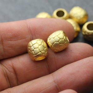 May include: Close-up of two gold-colored beads with a geometric pattern, resembling a soccer ball. The beads are cylindrical with a hole running through the center. Other similar beads are visible in the background.