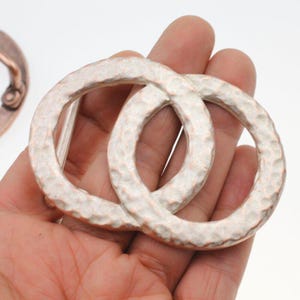 May include: Two silver-colored, hammered metal rings interlocked, held in a hand. The rings have a textured surface and a slightly oval shape. A copper-colored metal piece is visible in the background.