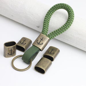 May include: Green rope keychain with a bronze-colored anchor emblem and matching metal accents. The keychain features a green leather strap and a bronze key ring. Several bronze metal pieces with anchor designs are scattered around the keychain.