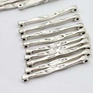 Puede incluir: Primer plano de varios componentes de joyería de metal plateado. Cada componente es una barra delgada y texturizada con un agujero en cada extremo. Los componentes tienen una apariencia martillada o desgastada, lo que sugiere un estilo vintage o artesanal.