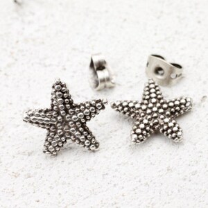 May include: A pair of silver starfish stud earrings. Each earring has a textured surface with small, round details. The earrings are shown with their backing posts and butterfly clasps, set against a white background.