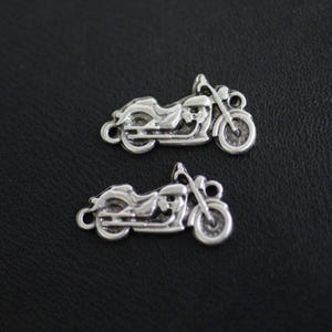 Könnte beinhalten: Zwei silberfarbene Motorrad-Anhänger. Jeder Anhänger zeigt ein detailliertes Motorraddesign mit Rädern, Lenker und Sitz. Die Anhänger sind klein und haben eine Schlaufe zum Befestigen an Schmuck oder anderen Bastelarbeiten. Der Hintergrund ist eine dunkle, strukturierte Oberfläche.