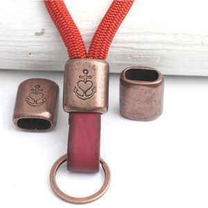 May include: A lanyard with a red cord and a burgundy leather strap, featuring antique bronze-colored metal accents. The metal pieces have an anchor and heart design. A bronze key ring is attached to the leather strap.