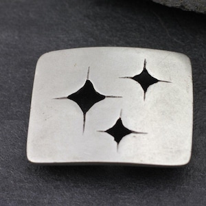 Puede incluir: Hebilla de cinturón de metal plateado con forma rectangular. La hebilla presenta tres recortes negros en forma de diamante, cada uno con cuatro líneas radiales. La hebilla tiene una superficie ligeramente texturizada y esquinas redondeadas.