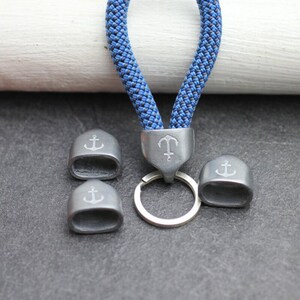 May include: A blue and black rope keychain with silver-colored metal accents. The keychain features a metal anchor charm and three additional metal pieces with anchor designs. The keychain is a nautical-themed accessory.