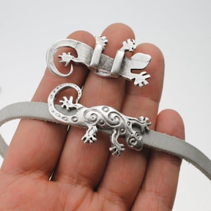May include: Two silver-colored lizard-shaped clasps attached to a light gray leather strap. The lizards have detailed designs and are positioned on a hand. The clasps are likely for a bracelet or necklace.