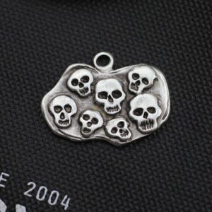 May include: A silver-toned pendant featuring seven detailed skull designs. The skulls are arranged on a rounded, irregular base with a small loop at the top for hanging. The pendant has a slightly aged appearance.
