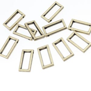 May include: A collection of rectangular metal charms in a bronze color. Each charm has a hollow, open frame design with small holes at the top and bottom. The charms are scattered on a white surface.