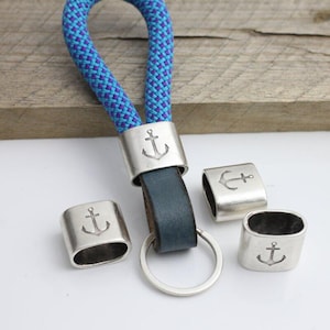 May include: A nautical-themed keychain with a blue and purple rope loop, a dark blue leather strap, and a silver-colored metal ring. The keychain features silver metal accents with an engraved anchor design.