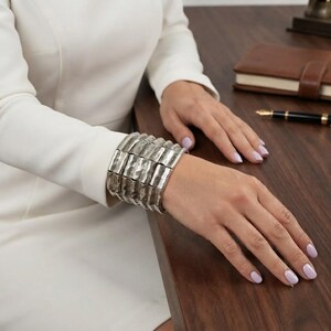 Könnte beinhalten: Ein breites, silberfarbenes Manschettenarmband bestehend aus einer Reihe verbundener, vertikal ausgerichteter rechteckiger Segmente mit einer texturierten, gehämmerten Oberfläche. Das Armband wird am Handgelenk getragen und zeigt einen metallischen Glanz.