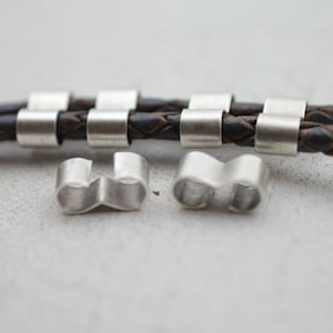 May include: Silver-colored metal bracelet clasps and connectors. The clasps are cylindrical and curved, designed to secure leather or braided cord bracelets. The background features a brown leather bracelet.