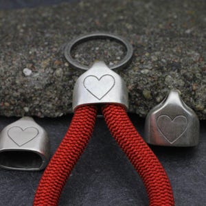 May include: A close-up of a red rope with silver-colored metal accents. The metal pieces have a heart design. The rope is attached to a metal ring. The background is a textured gray surface.