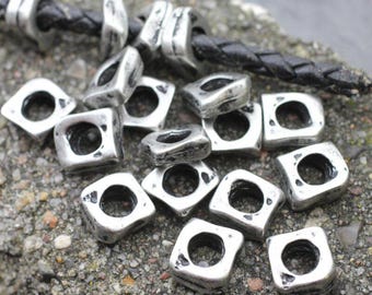May include: Silver-toned metal beads with a square shape and a central hole. The beads have a distressed, antique finish and are scattered on a textured surface. A black leather cord is visible in the background, with several beads strung on it.