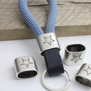 May include: A keychain with a blue and white rope loop, a black leather strap, and a silver-colored ring. The keychain has silver-colored metal accents with star designs. Several additional star-adorned metal pieces are scattered around the keychain.