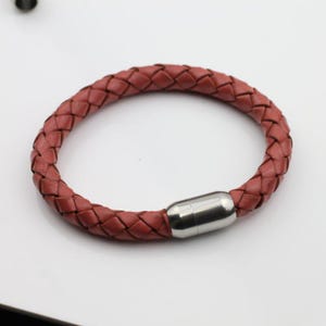 May include: A braided leather bracelet in a reddish-brown hue. The bracelet features a magnetic clasp made of polished silver-colored metal. The woven design gives the bracelet a textured appearance. This accessory is suitable for everyday wear.