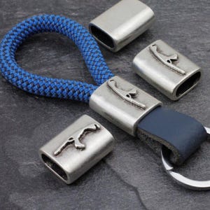 May include: A blue rope keychain with silver-colored metal accents. The keychain features a blue leather strap and a silver-colored metal clasp with a decorative design. Several additional metal pieces are scattered around the keychain.