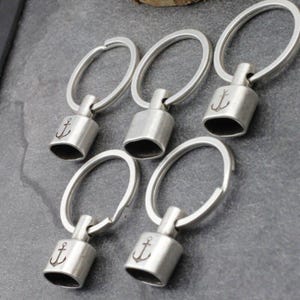 May include: Five silver-colored keychains with oval rings and cylindrical charms. Each charm features an engraved anchor symbol. The keychains are arranged on a gray surface, showcasing a nautical theme.