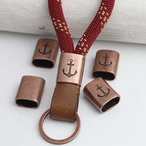 May include: A lanyard with a red and cream-colored rope design, a brown leather strap, and a copper-colored ring. Copper-colored metal pieces with anchor designs are scattered around the lanyard.