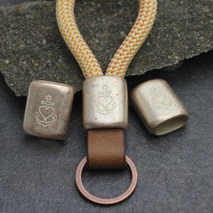May include: A keychain with a nautical theme. The design features three silver-colored metal beads with an anchor and heart emblem. The keychain includes a brown leather strap and a copper-colored ring. The rope is yellow and white.