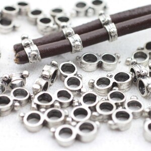 Pode incluir: Contas de charme de metal prateado em forma de óculos, espalhadas sobre uma superfície branca. Alguns encantos são presos a um cordão de couro marrom. Os encantos têm uma aparência vintage ou antiga.