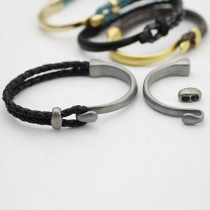 May include: A collection of bracelets with various designs. The foreground features a black braided leather bracelet with a silver-tone metal clasp. Other bracelets in the background have gold and black leather straps.
