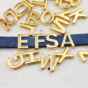Op de afbeelding: Goudkleurige metalen alfabetletters verspreid rond een blauwe leren band. De letters zijn in verschillende lettertypen en maten, met sommige letters die "EFSA" spellen. De letters zijn glanzend en reflecteren licht.