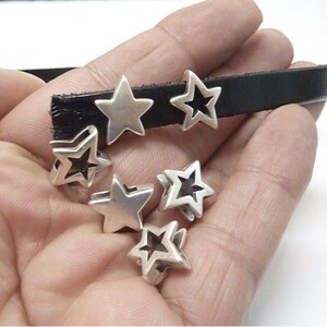 Può includere: Perline argentate a forma di stella, alcune con centri aperti, sono esposte su un cinturino in pelle nera. Le perline sono piccole e metalliche, con un design semplice e moderno. L'immagine mostra componenti di gioielleria.