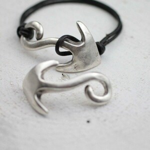 Puede incluir: Pulsera de ancla plateada con una banda de cuero negro. La pulsera presenta un diseño náutico con forma de ancla curvada. Un dije de ancla está suelto y acostado junto a la pulsera.