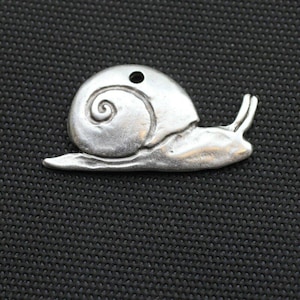 Pode incluir: Um berloque de caracol prateado com uma concha detalhada e um pequeno orifício para enfiar. O caracol é retratado em perfil, com o corpo estendido ao longo de uma base plana. O berloque é feito de metal.