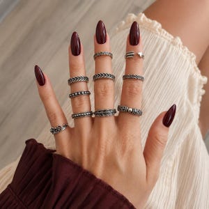 Può includere: Una collezione di dieci anelli in metallo color argento indossati sulle dita. Gli anelli presentano varie texture e motivi, inclusi design intrecciati, a perline, a freccia e a foglia. Ogni anello ha una finitura ossidata dettagliata.