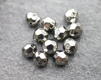 Könnte beinhalten: Eine Ansammlung silberfarbener, facettierter Perlen. Jede Perle hat eine vielseitige geometrische Form und einen metallischen Glanz. Die Perlen sind auf einer neutralen Oberfläche verstreut, was darauf hindeutet, dass sie für die Schmuckherstellung oder Bastelarbeiten bestimmt sind.