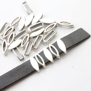 May include: Silver-toned oval and rectangular metal jewelry findings scattered on a white surface. Several oval findings are attached to a black leather strap, showcasing their potential use in jewelry design. The findings are bright and reflective.