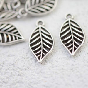 May include: Close-up of several silver-colored leaf charms. Each charm has a detailed leaf vein design and a small loop at the top for attaching to jewelry or other crafts. The charms are on a textured, light-colored surface.