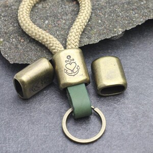 May include: A keychain with a tan rope loop and a green leather strap. The metal hardware is antique brass, including a rectangular piece with an anchor and heart design. Two additional metal pieces are included.