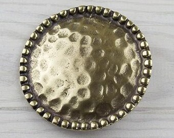 May include: A round, antique brass-colored decorative plate with a hammered texture and a beaded edge. The plate has a slightly concave center and is set against a light wood grain background.