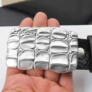 May include: A silver-coloured belt buckle with a rectangular shape and a textured, crocodile-skin design. The buckle is attached to a black leather belt. The buckle is held in a person's hand.