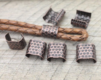 Könnte beinhalten: Kupferfarbene Metall-Crimp-Enden und Kordelenden mit gehammerter Textur und herzförmigen Aussparungen. Die Crimp-Enden sind zur Sicherung einer geflochtenen Lederschnur bestimmt. Die Komponenten werden auf einer hellen Holzoberfläche dargestellt.