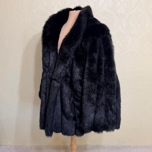 May include: A black faux fur coat featuring a wide shawl collar and a thick, plush texture. The garment has a straight, open-front design with vertical paneling throughout the body, providing a consistent, dense appearance across the entire piece.
