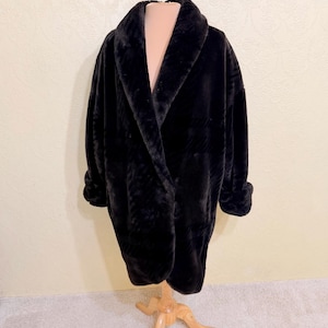 May include: A black faux fur coat featuring a wide shawl collar, long sleeves with cuffed ends, and a loose, open-front silhouette. The plush, textured material displays a subtle, tonal plaid pattern throughout the fabric of the garment.