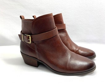 May include: A pair of brown leather ankle boots with a buckle strap. The boots have a low heel and a pointed toe.
