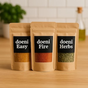Könnte beinhalten: Drei Gewürzbeutel aus Kraftpapier mit schwarzen Etiketten. Die Etiketten lauten "doeni Easy", "doeni Fire" und "doeni Herbs". Die Beutel enthalten gelbe, rote und grüne Gewürze. Auf einer Holzoberfläche.