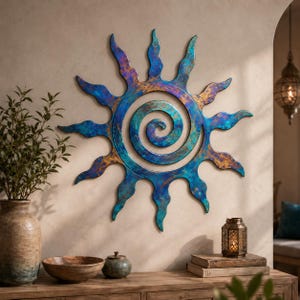 Könnte beinhalten: Ein kreisförmiges Wandkunstobjekt in Sonnenform mit einem zentralen Spiraldesign. Die Metalloberfläche weist ein gesprenkeltes Finish mit vermischten Tönen in Dunkelblau, Türkis und Metallic-Bronze auf, die in wellenförmigen Strahlen auslaufen.