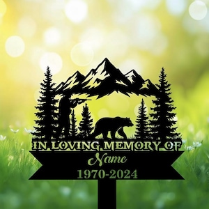 Könnte beinhalten: Schwarzer Metall-Gedenkpfahl mit Bergkette, Bäumen, einem Jäger und einem Bären. Der Text lautet "IN LOVING MEMORY OF" über einem Namen und den Daten 1970-2024. Der Hintergrund ist verschwommen grün und gelb.