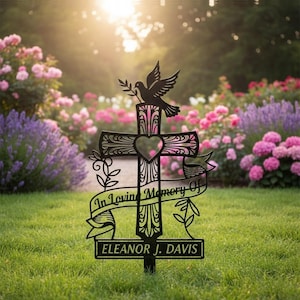 Könnte beinhalten: Schwarzes Metall-Gedenkkreuz mit einer Taube, die einen Olivenzweig hält. Das Kreuz hat einen herzförmigen Ausschnitt und Schnörkel, mit der Inschrift "In Loving Memory Of" und dem Namen "Eleanor J. Davis". In einem Blumengarten.