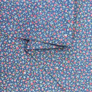 May include: A blue fabric featuring a dense, repeating pattern of small pink flowers with light green stems and tiny yellow dots scattered throughout. The material is folded, showing the texture and the consistent floral print design.