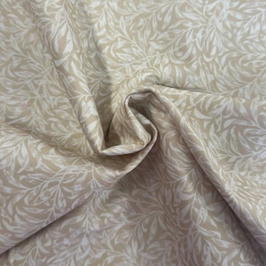May include: Beige fabric featuring a repeating pattern of white, stylized leaves. The fabric is folded, creating shadows and texture. The leaves have a delicate, flowing design, suitable for various sewing projects.