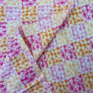 May include: A folded fabric featuring a gingham pattern in pink and orange, adorned with a delicate floral design. The textile combines bright and pastel hues, creating a cheerful and visually appealing fabric.
