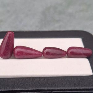 May include: Four polished, deep magenta-coloured teardrop-shaped gemstones arranged in a row. One stone stands vertically on its rounded end, while the other three lie horizontally. The stones have a smooth, glossy surface with subtle internal textures.