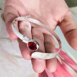 May include: A silver cuff bracelet featuring a large, oval, deep red gemstone. The bracelet has a smooth, polished finish and a simple design. The text "jbhjewel" is visible on the bracelet.