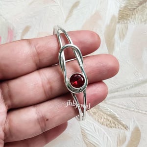 Könnte beinhalten: Ein silberfarbenes Armreif-Armband mit einem leuchtenden, runden, tiefroten Edelstein. Das Armband hat ein einzigartiges Design mit einer zentralen Schlaufe, die den Edelstein hält. Der Text "jhbjewel" ist sichtbar.